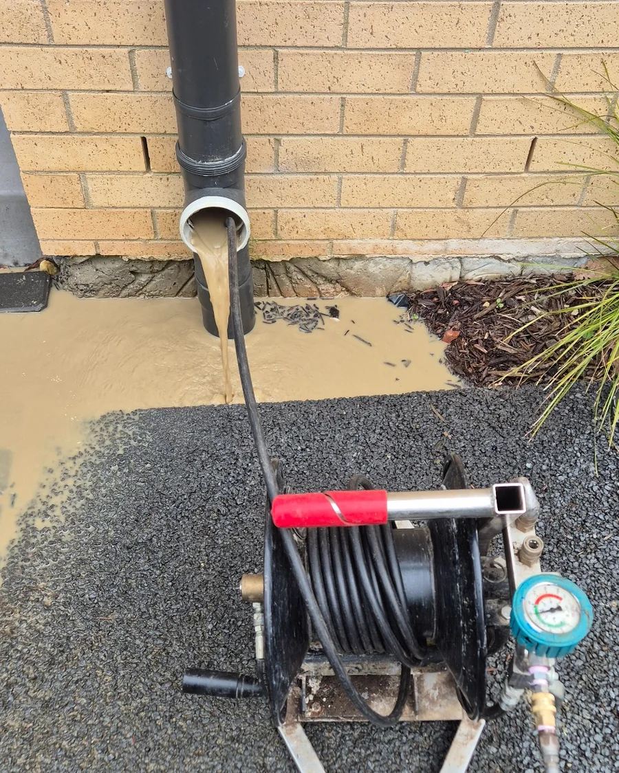 Blocked stormwater downpipe overflowing with muddy water while high-pressure hydro jetting equipment is used to clear the drain.