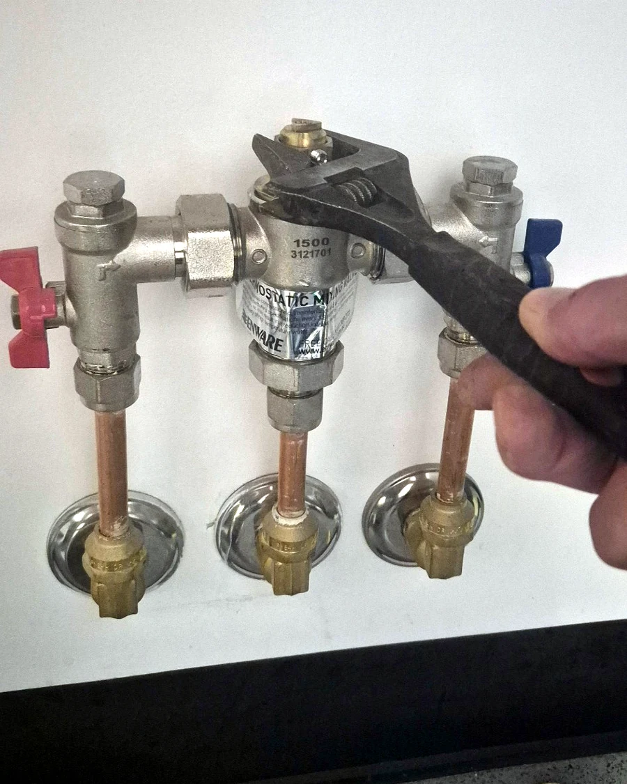 Plumber adjusting a tempering valve under a basin during hot water temperature control testing.
